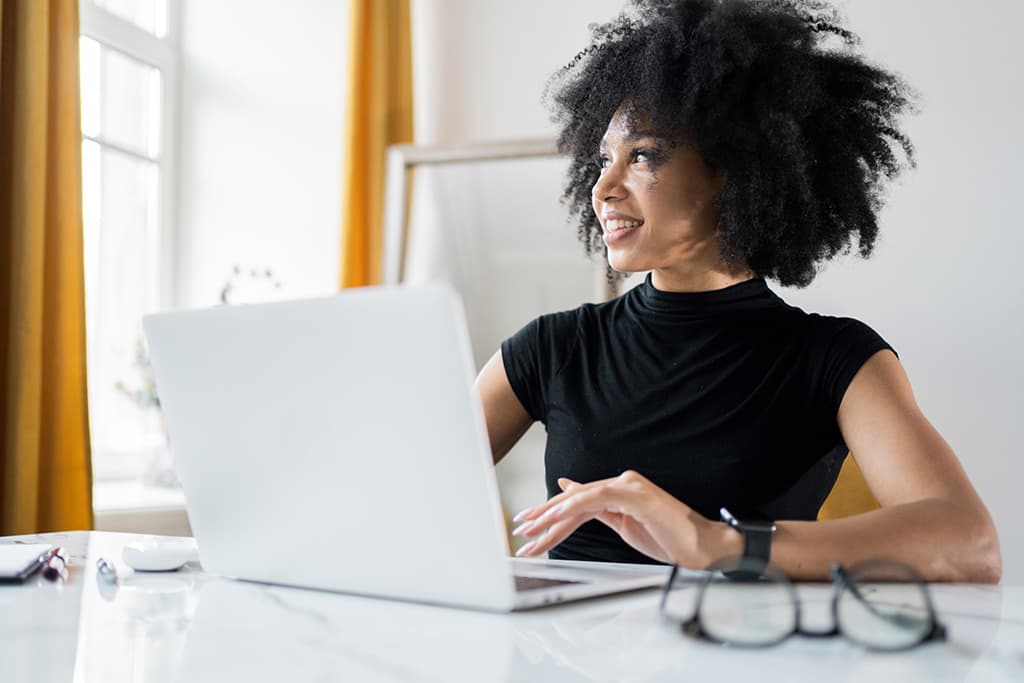 A woman working on her computer pausing to look out a bright window.